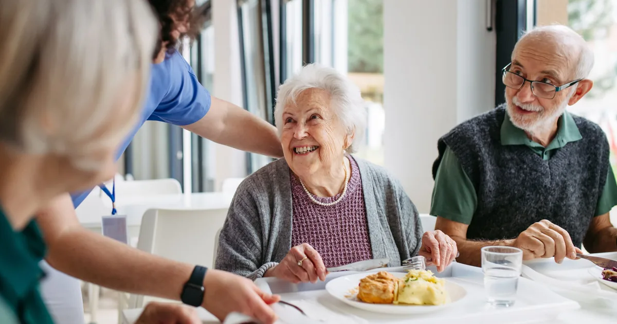 A carer chatting with residents over lunch in a care home dining room